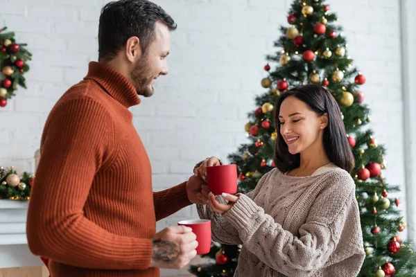 Homem sorrindo dando xícara de cacau quente para a esposa satisfeita perto da árvore de natal decorada — Fotografia de Stock