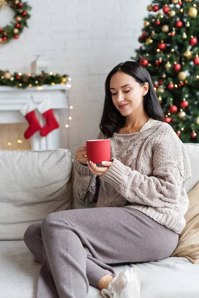 Задоволена брюнетка з чашкою какао сидить на дивані у вітальні з прикрашеною різдвяною ялинкою — стокове фото