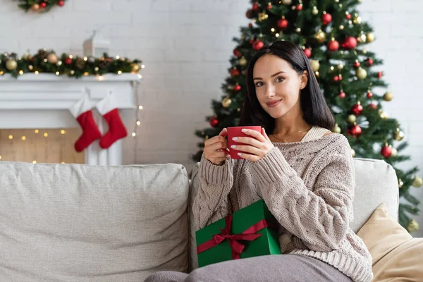 Glückliche Frau mit einer Tasse warmen Kakaos und Geschenkschachtel, die auf einer Couch im Wohnzimmer mit Weihnachtsdekor sitzt — Stockfoto