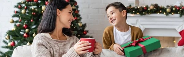 Mujer feliz con taza de cacao caliente mirando hijo alegre con regalo de Navidad, bandera - foto de stock