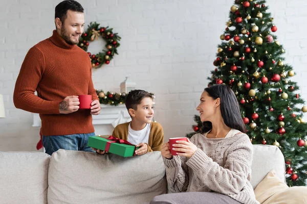 Menino feliz segurando Natal presente perto dos pais com copos de cacau quente — Fotografia de Stock