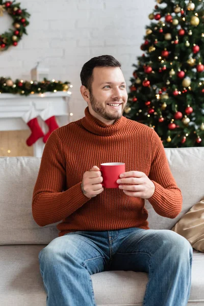 Glücklicher Mann mit Tasse Kakao auf Sofa in der Nähe des Weihnachtsbaums auf verschwommenem Hintergrund — Stockfoto