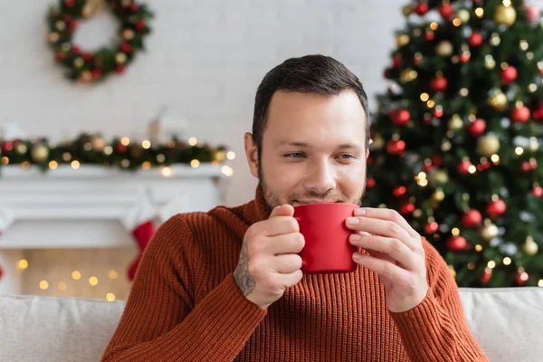 Fröhlicher Mann trinkt warmen Kakao in verschwommenem Wohnzimmer mit Weihnachtsdekor — Stockfoto