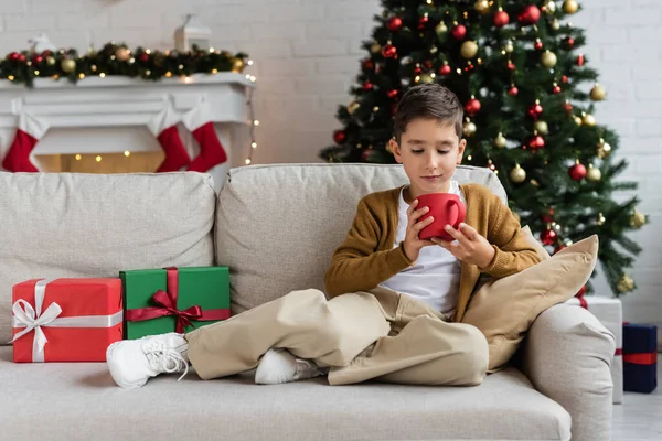 Piena lunghezza del capretto che tiene la tazza di cacao caldo sul divano vicino alle scatole regalo e all'albero di Natale offuscato — Foto stock