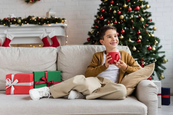 Menino alegre sentado no sofá com xícara de cacau e olhando para longe perto de caixas de presente e árvore de natal borrada — Fotografia de Stock