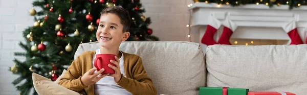 Niño feliz sentado en el sofá con la taza de cacao cerca del árbol de Navidad borrosa, pancarta - foto de stock