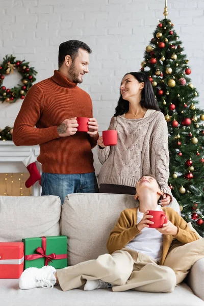Marido y mujer con tazas de cacao sonriéndose el uno al otro cerca del hijo en el sofá con regalos de Navidad - foto de stock