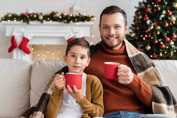 Feliz padre e hijo con tazas de cacao sentado debajo de una manta caliente en la sala de estar con una decoración de Navidad borrosa - foto de stock