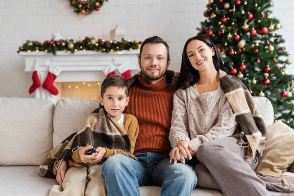 Menino com controlador remoto perto de família feliz assistindo tv no sofá sob cobertor quente perto de árvore de natal borrada — Fotografia de Stock