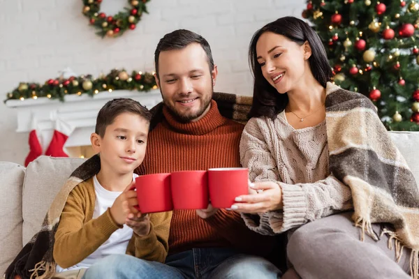 Cheerful family clinking cups with cocoa on couch under warm blanket near blurred christmas tree — Stock Photo