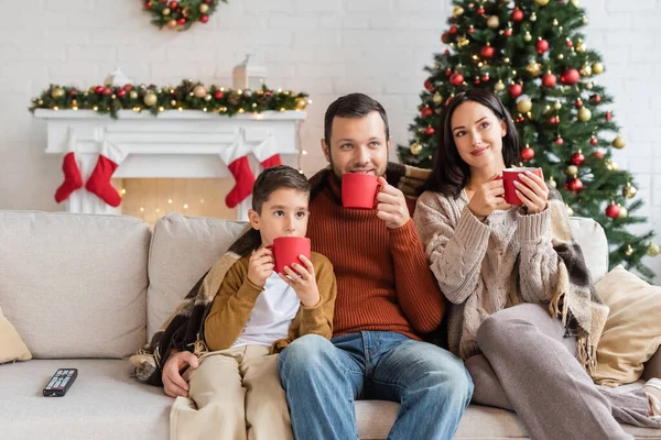 Familia feliz beber cacao en el sofá debajo de la manta cerca del árbol de navidad borrosa - foto de stock