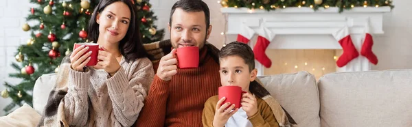 Familia feliz beber cacao bajo una manta caliente en la sala de estar con decoración de Navidad, pancarta - foto de stock