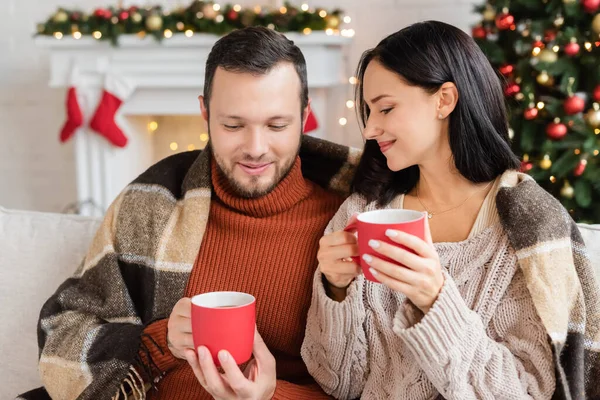Lächelndes Paar mit Tassen Kakao unter warmer Decke im Wohnzimmer mit verschwommenem Weihnachtsdekor — Stockfoto