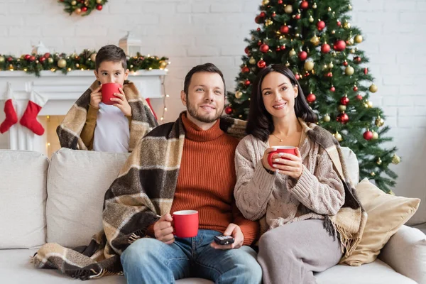 Familia sonriente con mantas calientes sosteniendo tazas de cacao en la sala de estar con decoración de Navidad - foto de stock