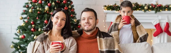 Cheerful couple sitting under warm blanket near son drinking cocoa at christmas tree, banner — Stock Photo