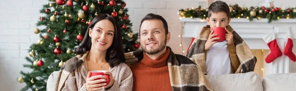 Couple joyeux assis sous une couverture chaude près de fils boire du cacao à l'arbre de Noël, bannière — Photo de stock