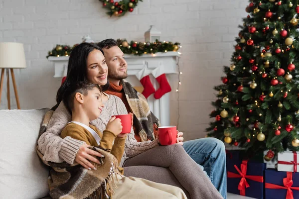 Joyeuse famille boire du cacao sur le canapé sous une couverture chaude près de boîtes-cadeaux sous l'arbre de Noël décoré — Photo de stock