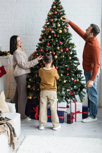 Семейное украшение елки рождественскими шарами возле подарочных коробок в гостиной — стоковое фото