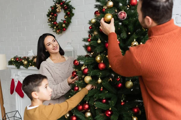 Mulher morena alegre olhando para marido e filho decorando árvore de Natal na sala de estar — Fotografia de Stock