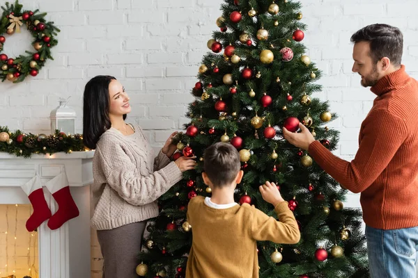 Cheerful woman decorating green spruce with baubles near family and fireplace with christmas stockings — Stock Photo