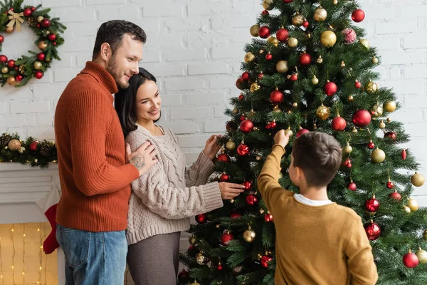 Homem sorridente abraçando alegre esposa decorando abeto verde com bugigangas de Natal perto do filho — Fotografia de Stock