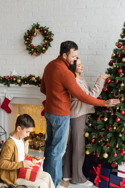Junge packt Weihnachtsgeschenk ein, während Eltern grüne Fichte zu Hause mit Kugeln dekorieren — Stockfoto