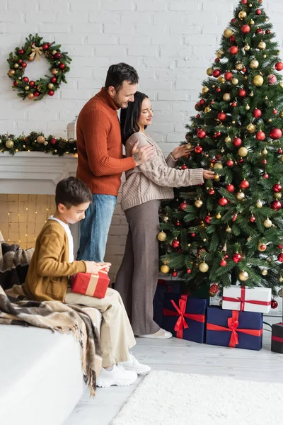 Vue latérale de souriant homme étreignant femme décoration arbre de Noël près de fils emballage boîte cadeau sur canapé — Photo de stock