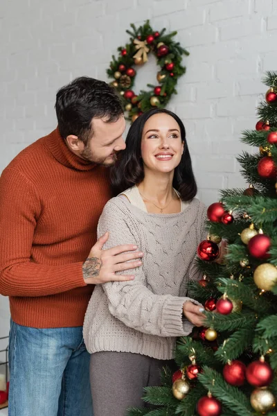 Мужчина обнимает весёлую брюнетку жена украшает рождественскую ёлку дома — стоковое фото