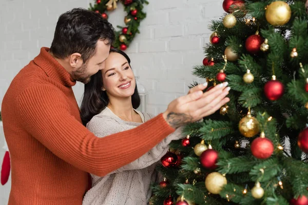 Fröhliches Paar dekoriert grüne Fichte mit Weihnachtskugeln im Wohnzimmer — Stockfoto