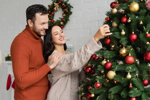 Uomo sorridente abbracciando felice e bella moglie decorare abete rosso verde con le bagattelle di Natale — Foto stock