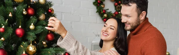 Glücklicher Mann lächelt neben brünetter Frau, die grüne Kiefer mit Weihnachtskugeln dekoriert, Banner — Stockfoto