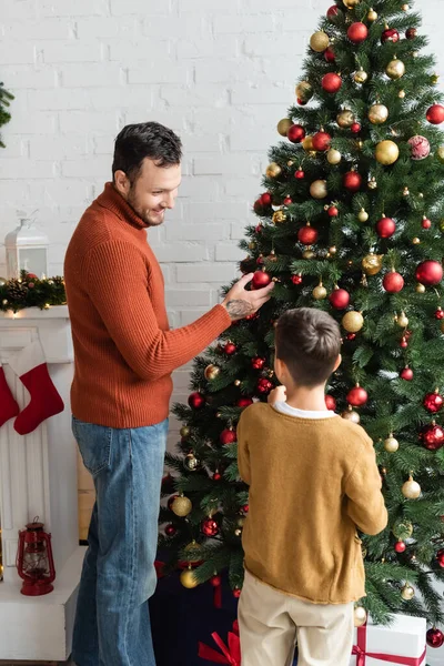 Lächelnder Mann schaut Sohn beim Schmücken des Weihnachtsbaums zu Hause mit Kugeln an — Stockfoto