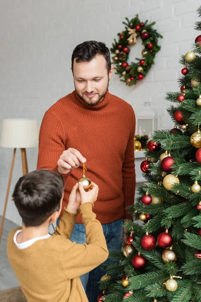 Lächelnder Papa übergibt Weihnachtskugel an Sohn, der grüne Fichten zu Hause dekoriert — Stockfoto