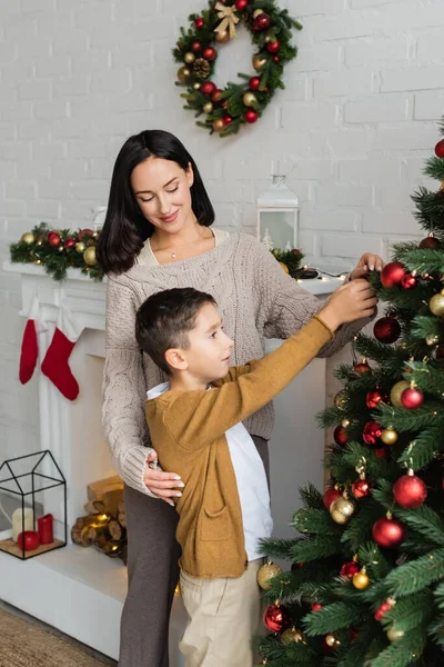 Sonriente mujer ayudando a su hijo a decorar pino verde con adornos de Navidad cerca de la chimenea en casa - foto de stock