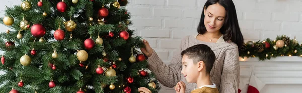 Brünette Frau lächelt neben Sohn, der grüne Fichte mit Weihnachtskugeln dekoriert, Banner — Stockfoto