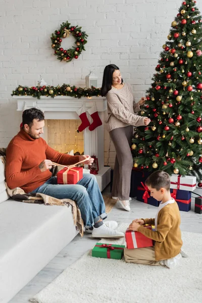 Happy woman decorating christmas tree near father and son packing presents in living room with fireplace — Stock Photo