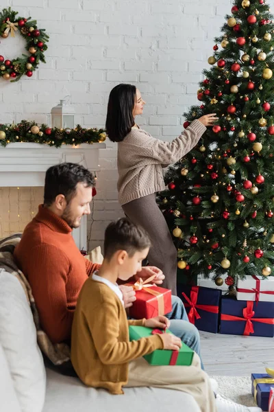 Vue latérale de la femme décorant l'arbre de Noël près de mari et fils emballant des cadeaux sur le canapé — Photo de stock