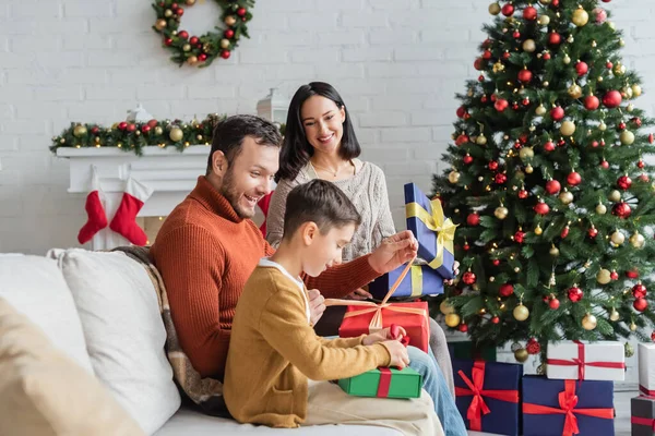 Alegres cajas de regalo de embalaje familiar en el sofá cerca del árbol de Navidad y chimenea decorada - foto de stock