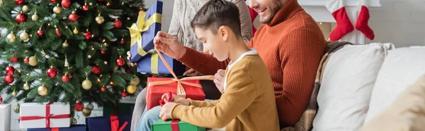 Criança embalagem caixa de presente perto de pais e árvore de natal em casa, banner — Fotografia de Stock