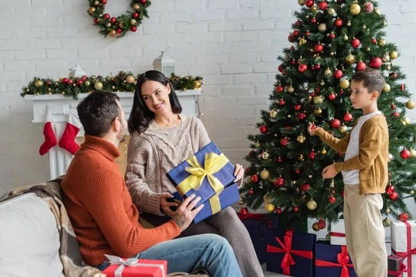 Niño decorando árbol de Navidad cerca de padres felices con caja de regalo sonriendo el uno al otro en el sofá - foto de stock