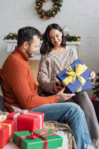 Smiling woman holding christmas present while sitting on couch near husband and gift boxes — Stock Photo