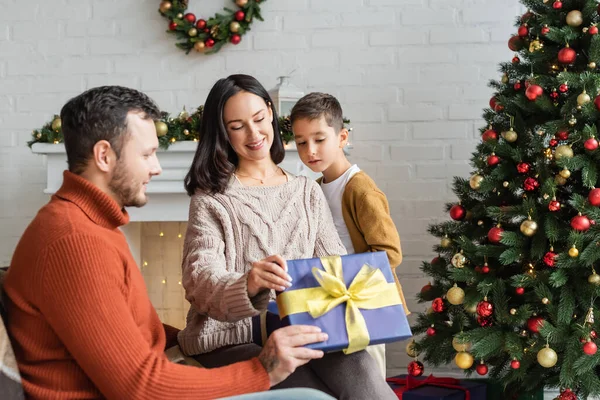 Happy woman holding gif box near husband and son in living room with decorated christmas tree — Stock Photo