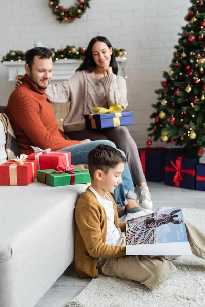 Menino sorrindo leitura revista no chão perto de pais desfocados e presentes de Natal no sofá na sala de estar — Fotografia de Stock