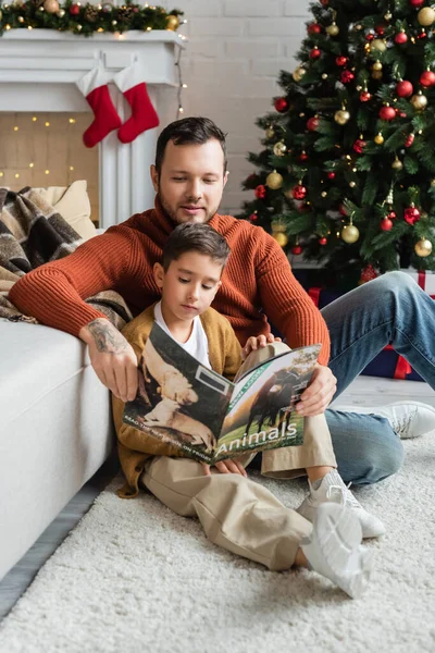 Pai e filho sentado no chão na sala de estar e revista de leitura perto da árvore de natal — Fotografia de Stock