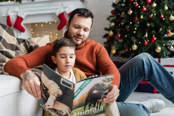 Menino leitura revista com sorrindo pai perto borrada árvore de Natal — Fotografia de Stock