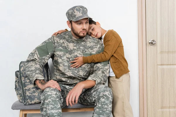 Kid with closed eyes embracing serious dad in camouflage sitting near backpack — Stock Photo