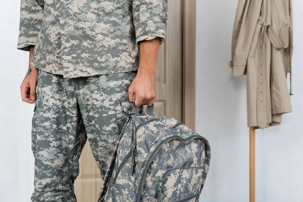 Vista cortada de soldado em camuflagem em pé com mochila em casa — Fotografia de Stock