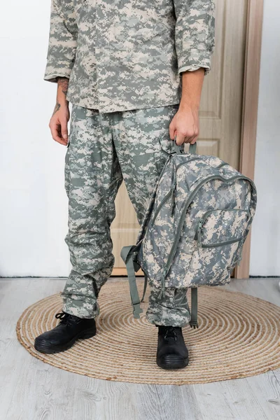 Vue partielle du militaire en uniforme et bottes debout avec sac à dos près de la porte d'entrée à la maison — Photo de stock