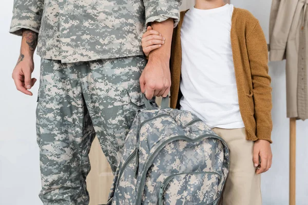 Частковий вид військовослужбовця з рюкзаком біля сина тримає руку вдома — стокове фото