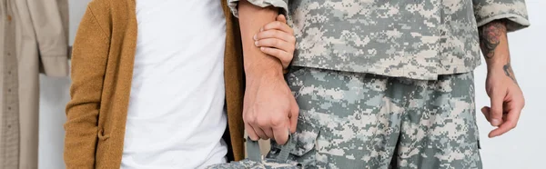 Vista parcial del niño cogido de la mano del padre en camuflaje mientras se reunía con él en casa, pancarta - foto de stock